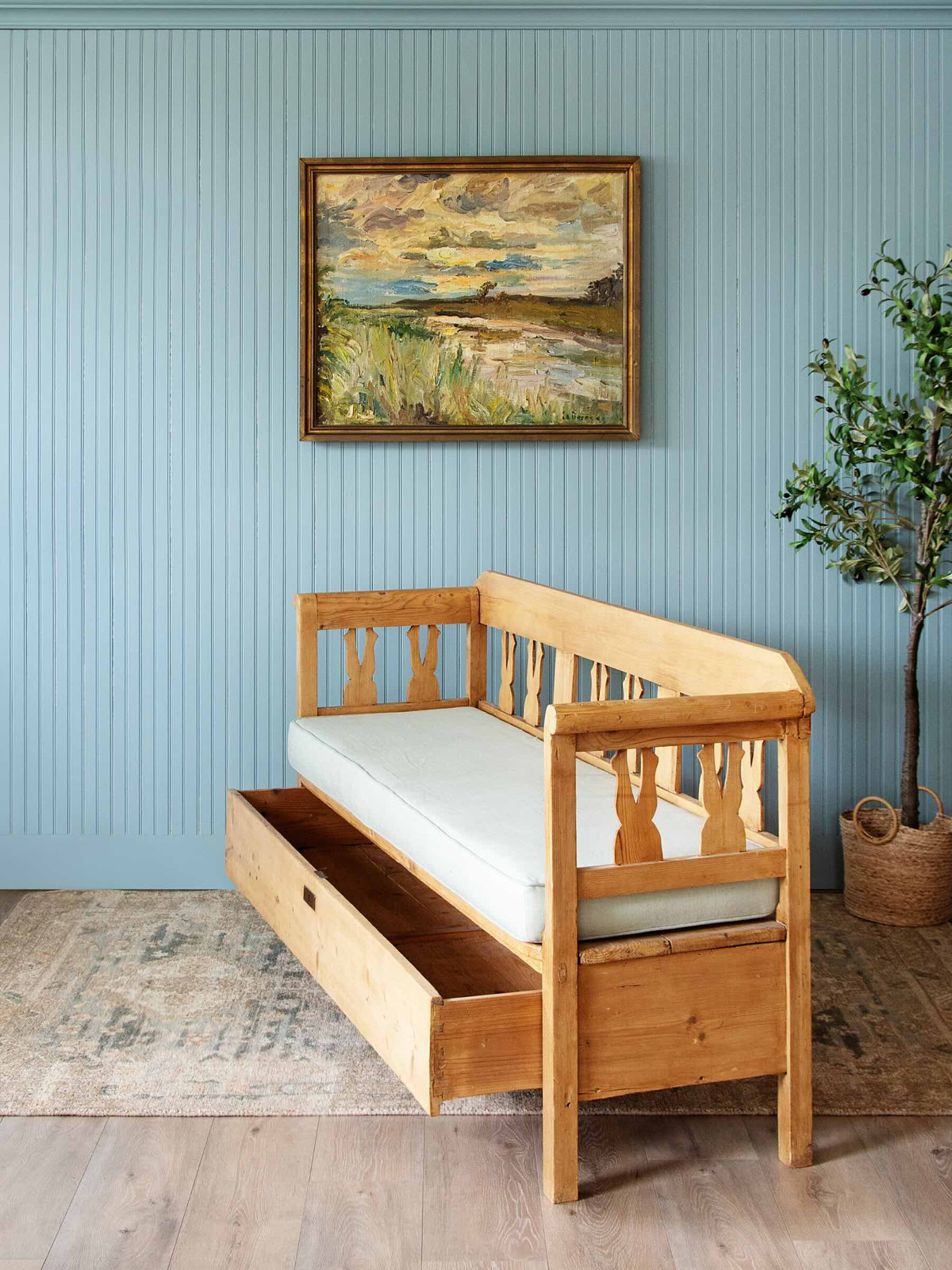 A light natural wood daybed with a white mattress, featuring a pull-out storage drawer underneath, set against a light blue paneled wall with a framed landscape painting and a potted plant.