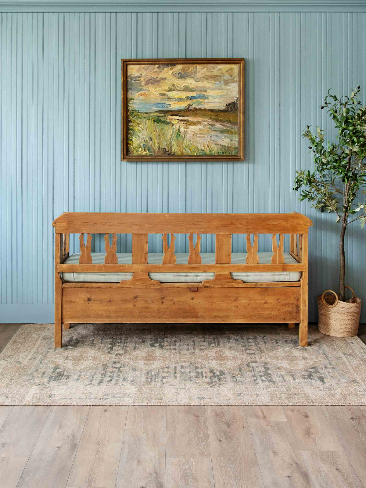 A rustic wooden daybed with a slatted back and seat, styled in a bedroom setting against a light blue paneled wall. A framed landscape painting hangs above the bed, and a potted tree stands on a patterned rug next to it.