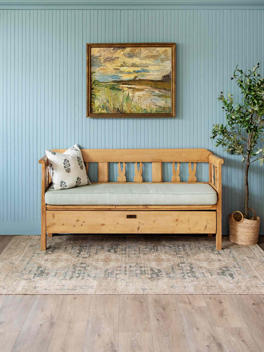 A light natural wood daybed with a pale green cushion and a patterned throw pillow, set against a light blue paneled wall with a framed landscape painting and a potted tree.