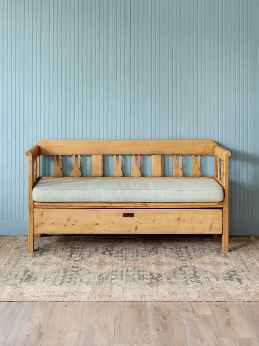 A light natural wood daybed with a ribbed slatted backrest and a pale sage green cushion, set against a light blue paneled wall on a patterned rug.