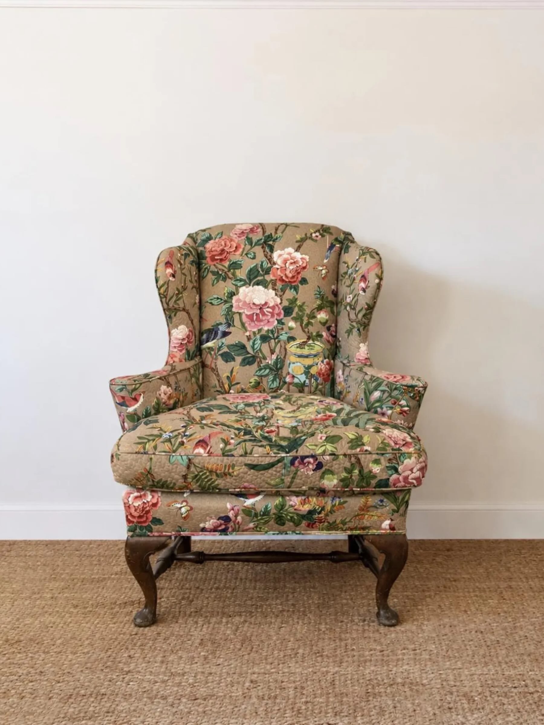 A classic wingback armchair upholstered in a vintage floral and bird patterned fabric, featuring a beige background with pink roses, green leaves, and small birds, supported by dark wooden cabriole legs, set against a plain white wall and light brown woven rug.
