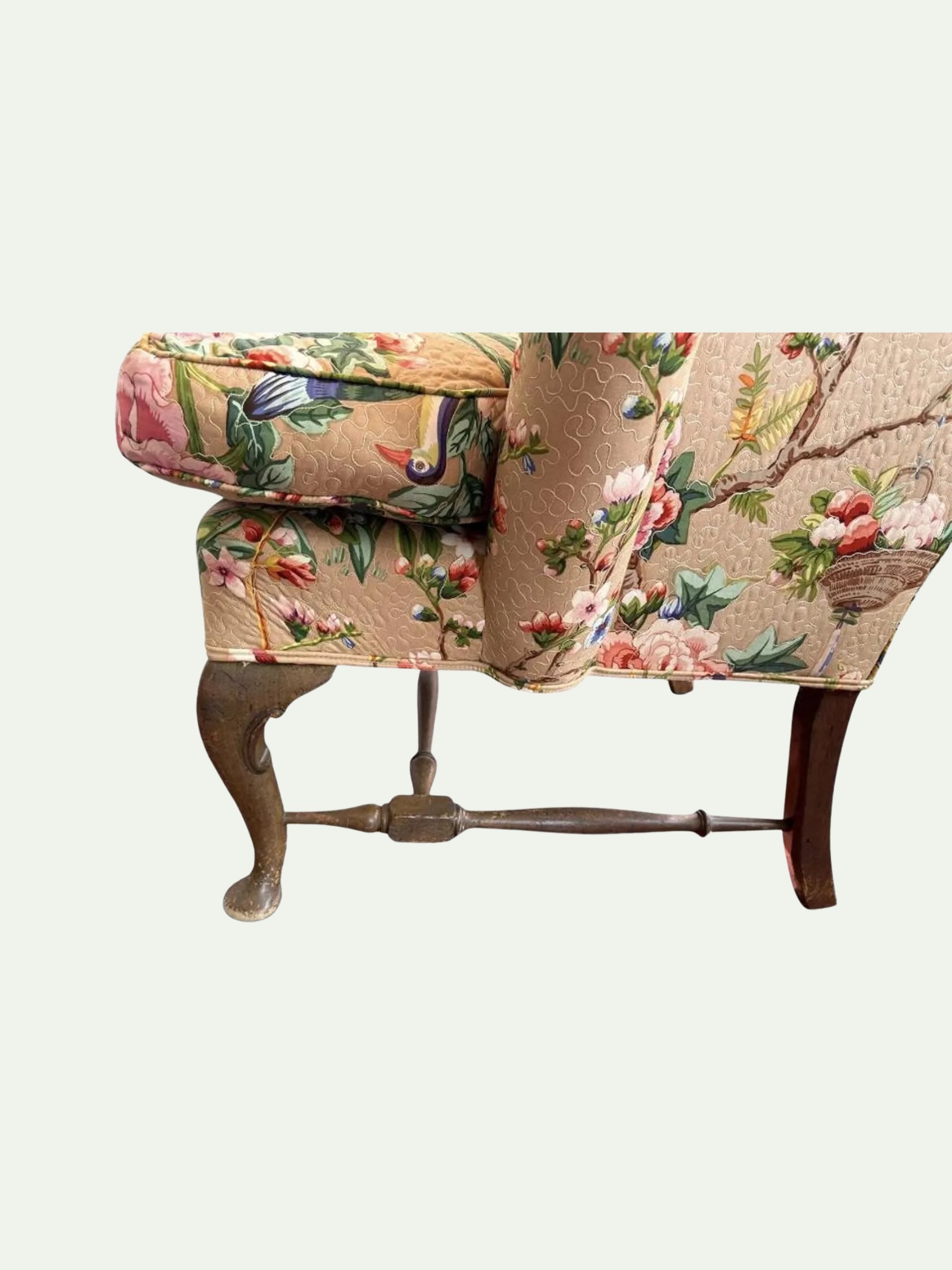 A close-up of an upholstered armchair or bench section showing the back and side, covered in a beige fabric with a colorful floral and bird print, supported by dark wooden legs and a central metal crossbar.