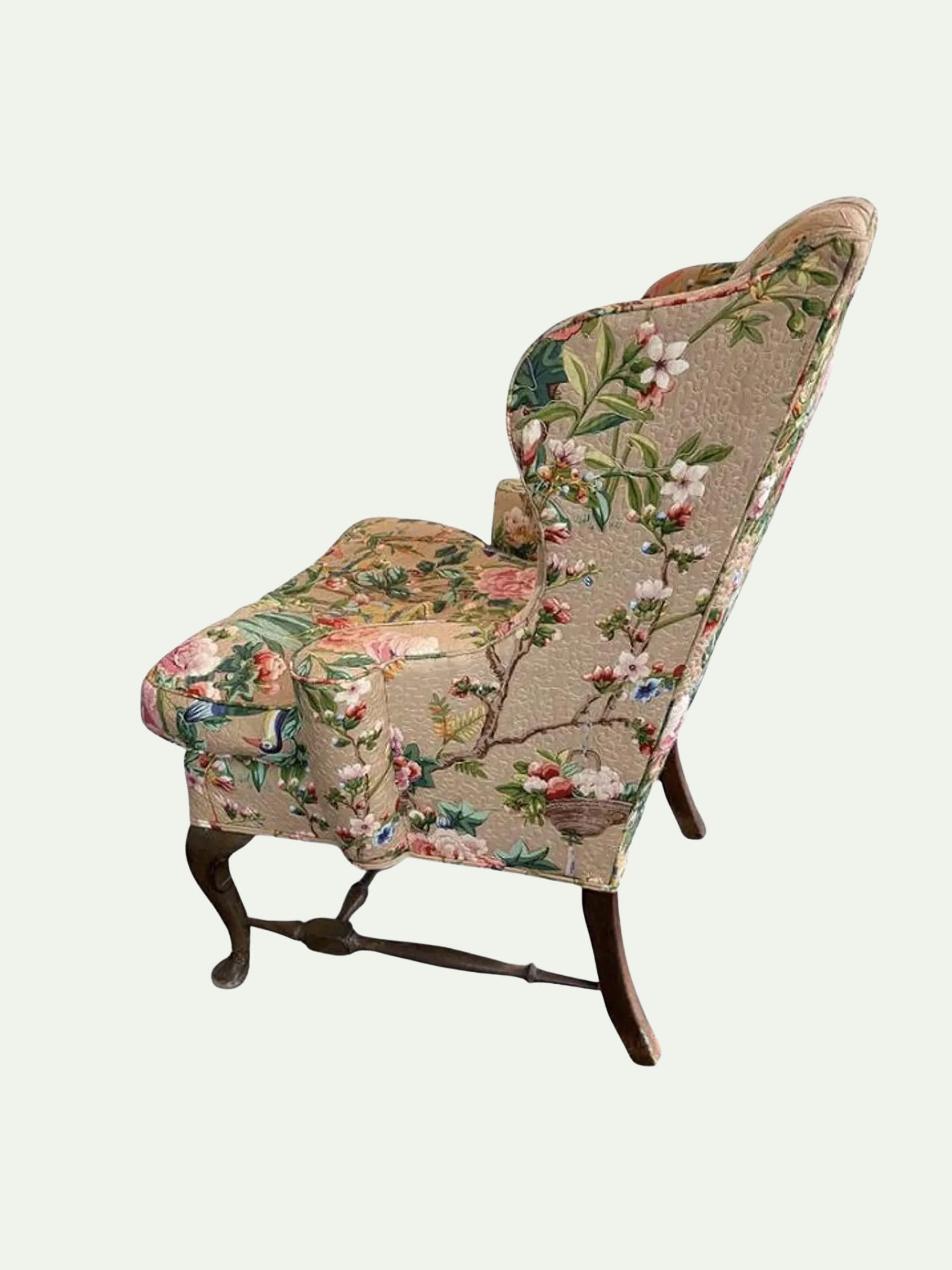 A vintage-style wingback armchair upholstered in a beige floral tapestry fabric with pink, green, and blue botanical motifs, supported by dark wooden legs.