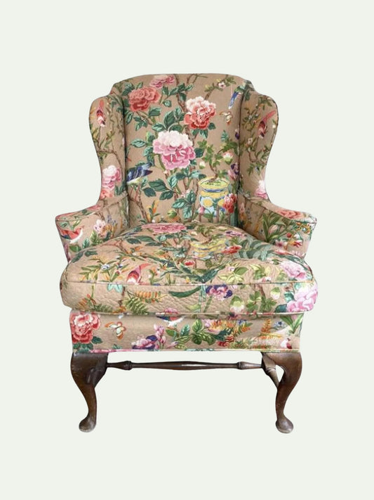 A classic wingback armchair upholstered in a detailed floral and bird patterned fabric with dark brown wooden cabriole legs, shown against a solid light gray background.