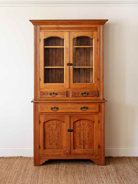 A tall wooden hutch cabinet with a warm medium-brown finish, featuring two upper glass-front doors, two lower doors with decorative metal floral motifs, and a single central drawer. The cabinet stands on a woven jute rug against a white wall.