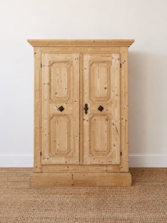 A light natural wood double-door cabinet with distressed black metal handles, standing on a woven jute rug against a plain off-white wall.
