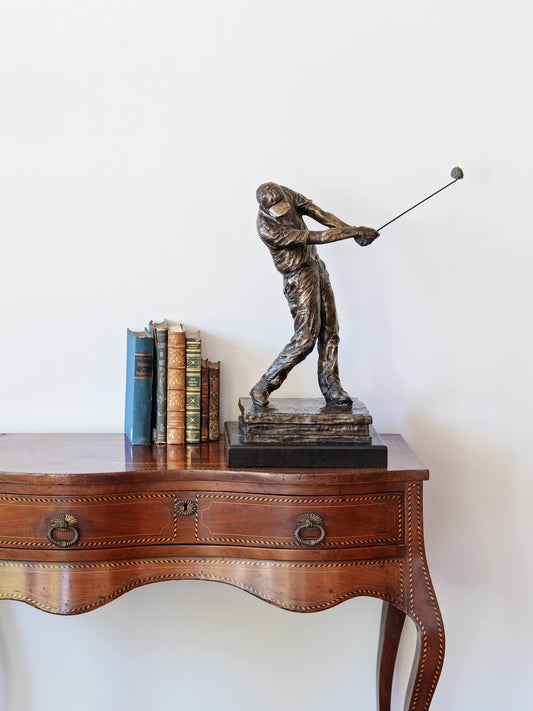 A bronze-finished sculpture of a golfer mid-swing, displayed on a dark wooden console table next to a set of books against a plain white wall.