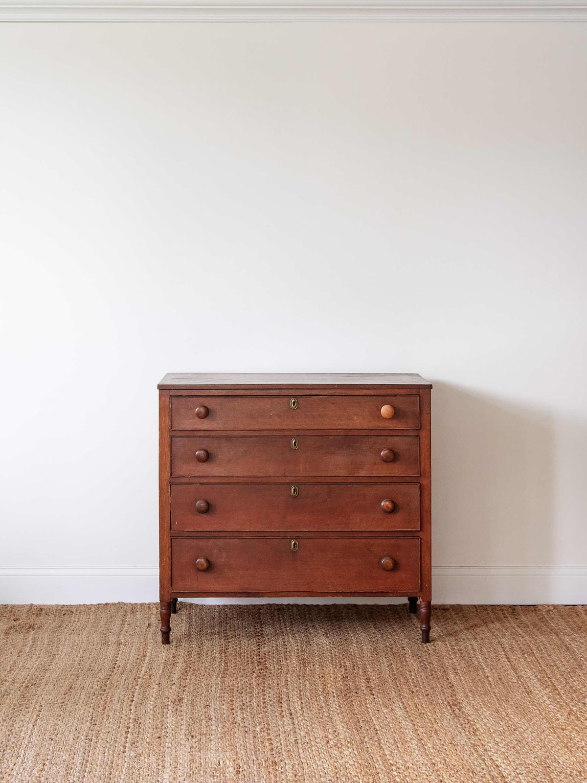 A vintage four-drawer wooden chest of drawers with round knobs and brass hardware, set against a plain white wall on a woven jute rug.