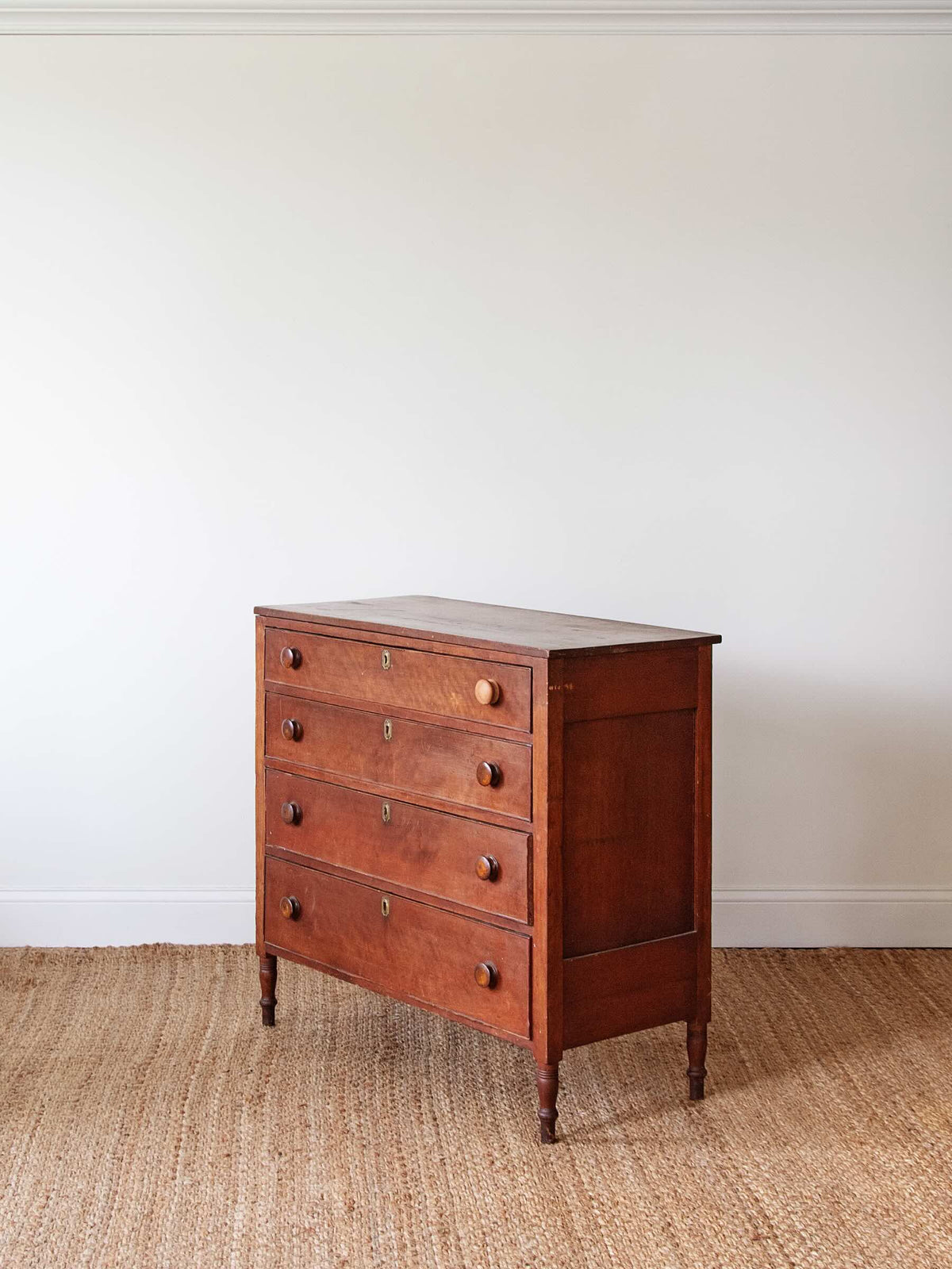 A vintage wooden chest of drawers with five drawers and small round knobs, standing on a woven jute rug against a plain off-white wall.
