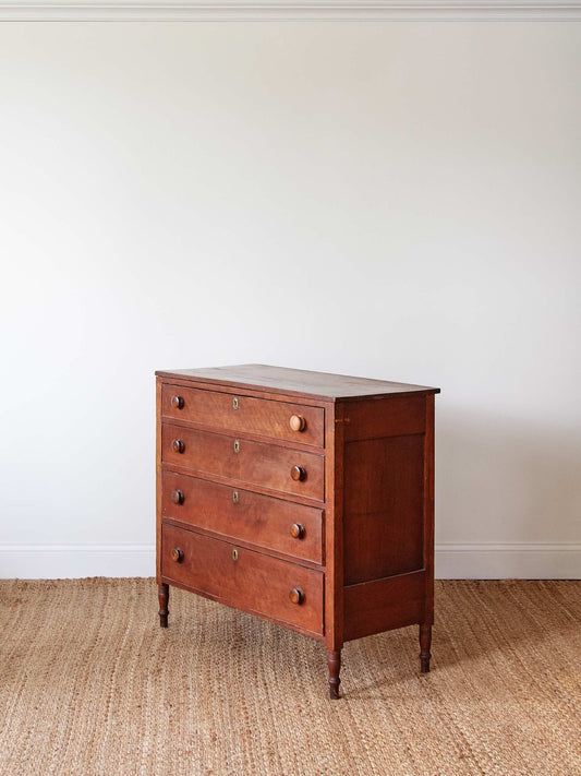 A vintage wooden chest of drawers with five drawers and small round knobs, standing on a woven jute rug against a plain off-white wall.