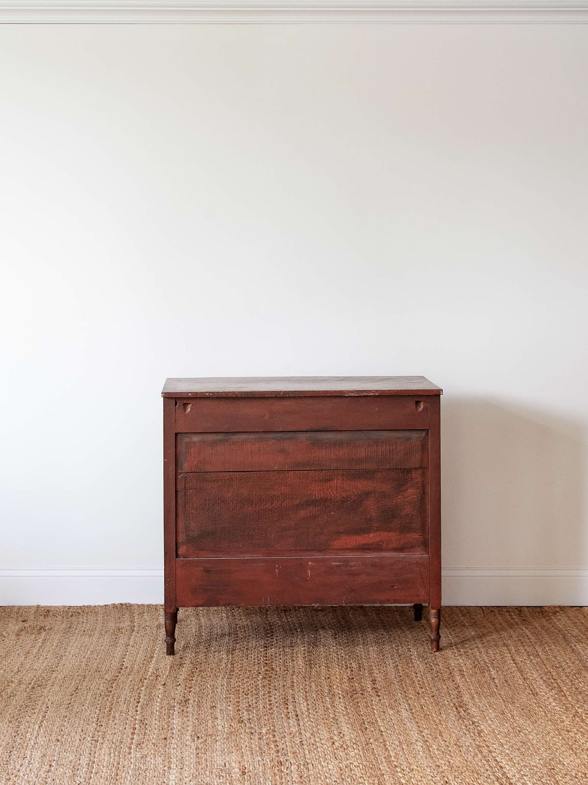 A vintage reddish-brown wooden chest or cabinet with a single drawer, standing on a woven jute rug against a plain white wall.