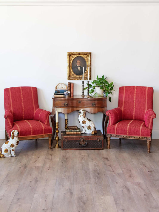 A styled interior scene with two red upholstered armchairs flanking a wooden console table against a white wall. Decorative items on the table include books, a candlestick, a small plant, and a framed portrait.