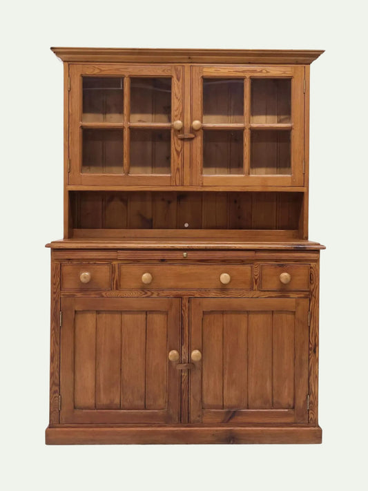 A rustic wooden hutch cabinet with a warm medium-brown finish, featuring a glass-front upper cabinet, an open shelf, three drawers, and two lower cabinet doors with round wooden knobs, set against a plain off-white background.