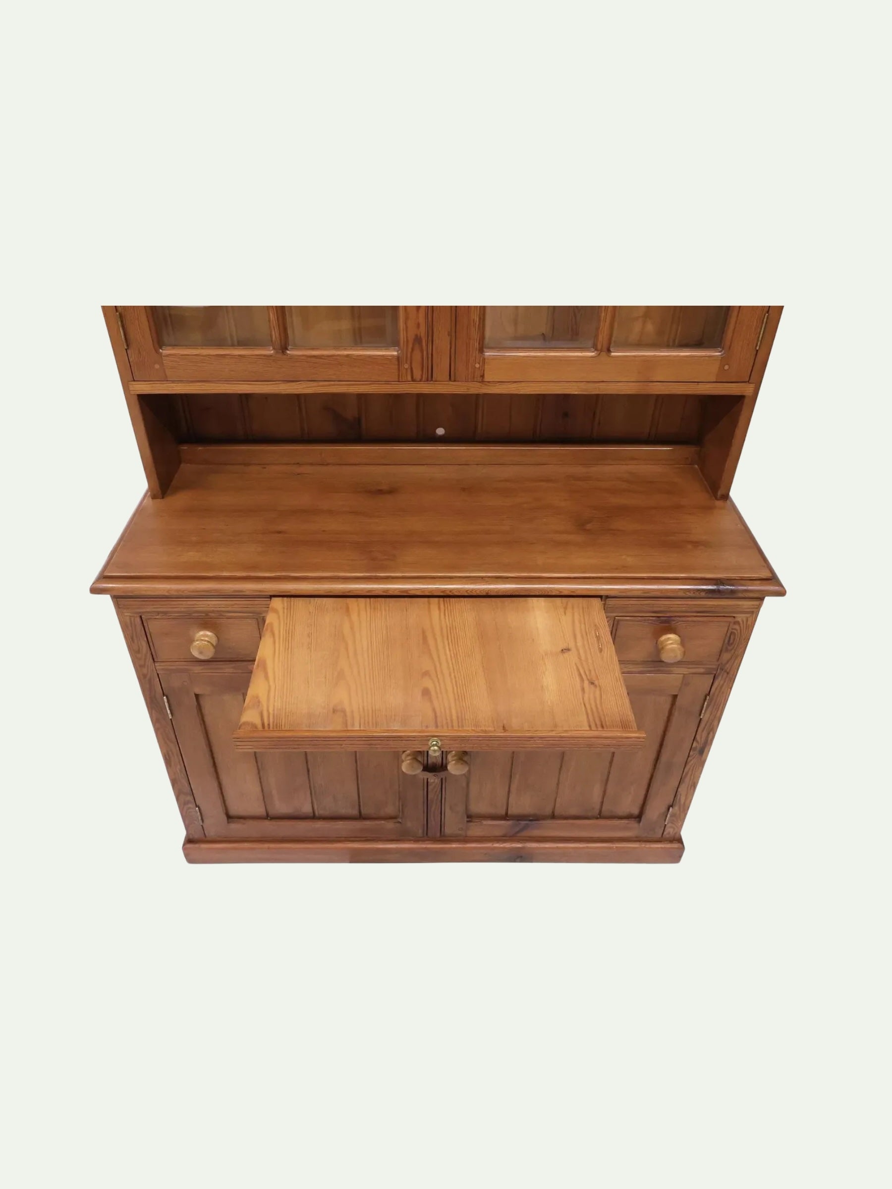 A mid-century modern wooden credenza or hutch with a warm medium-brown finish, featuring a pull-out writing desk, two drawers with round knobs, and a glass-front upper cabinet section, shown against a plain off-white background.