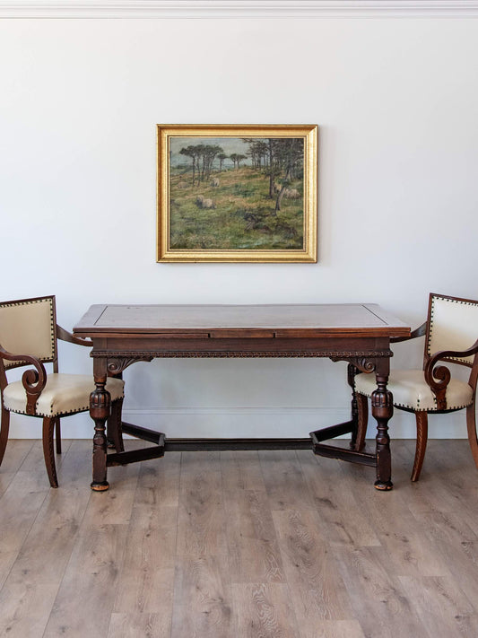A dark wood rectangular dining table with carved legs is shown in a room setting, flanked by two cream upholstered wooden chairs. A framed landscape painting hangs on the white wall above the table, and light wood flooring is visible.