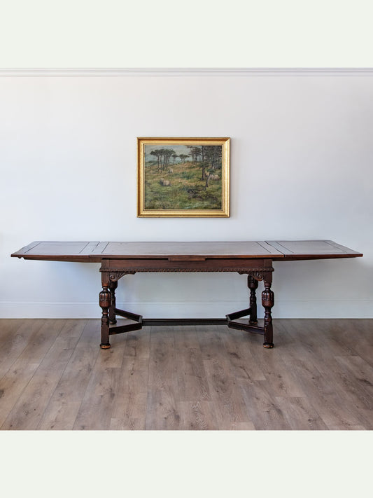 A dark wood extendable dining table with turned legs and stretcher supports, set against a white wall with a framed landscape painting above it and light wood flooring.