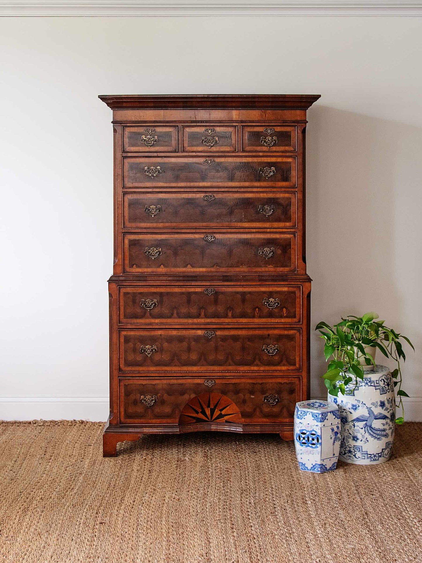 A tall, dark brown wooden chest of drawers with multiple drawers and ornate metal pulls, set against a plain white wall on a woven jute rug. Two blue and white ceramic garden pots with green plants sit to the right of the dresser.