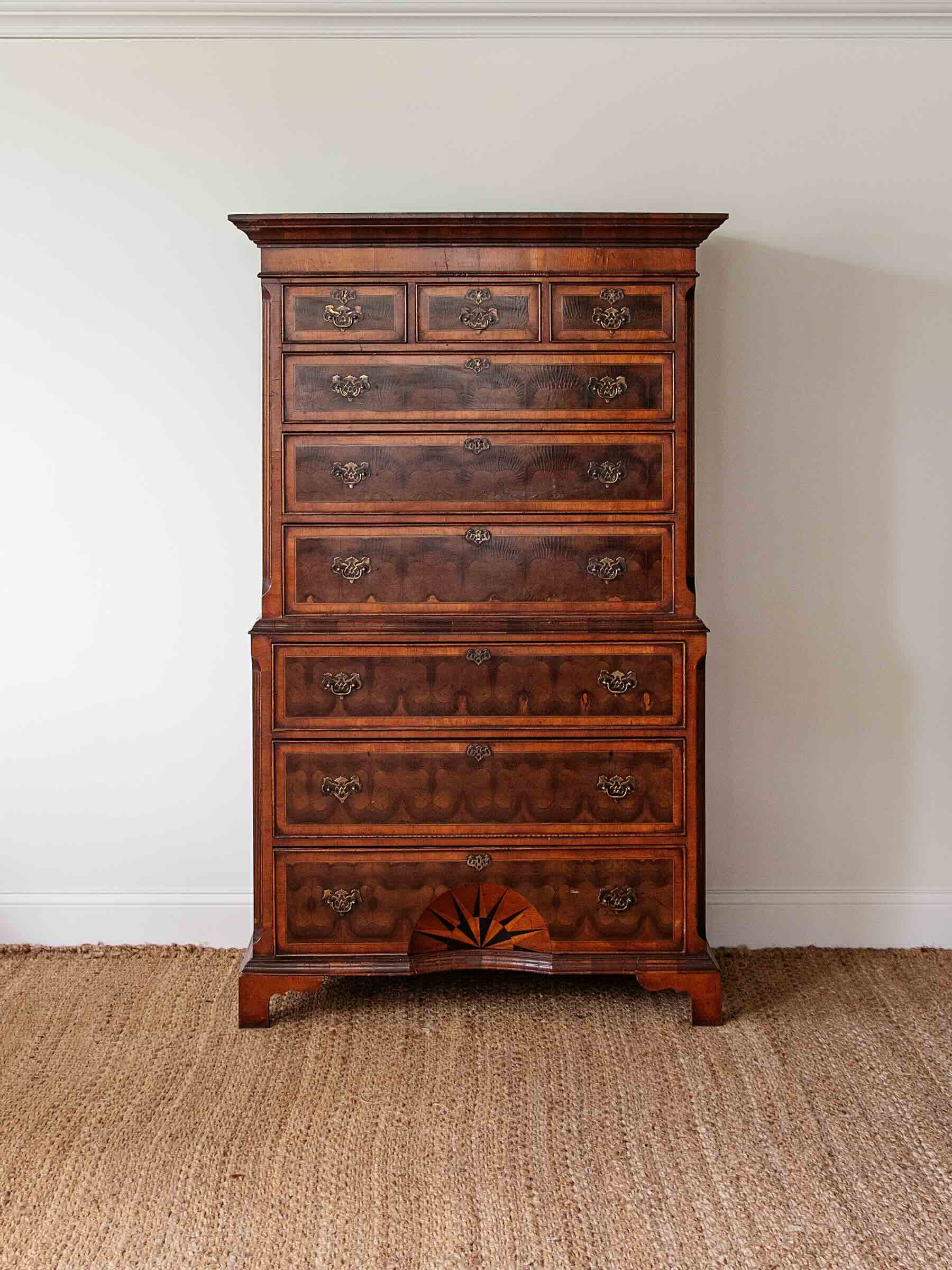 A tall, dark brown wooden chest of drawers with multiple drawers and ornate metal pulls, set against a plain white wall on a woven jute rug.