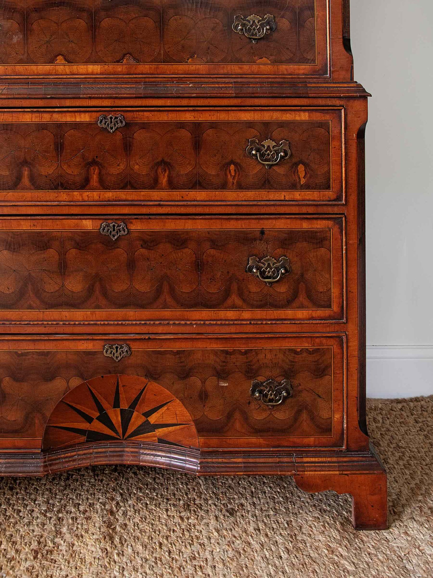 Close-up of an antique wooden chest of drawers with intricate marquetry patterns, brass drawer pulls, and a star inlay on the lower drawer, set on a woven rug against a white wall.