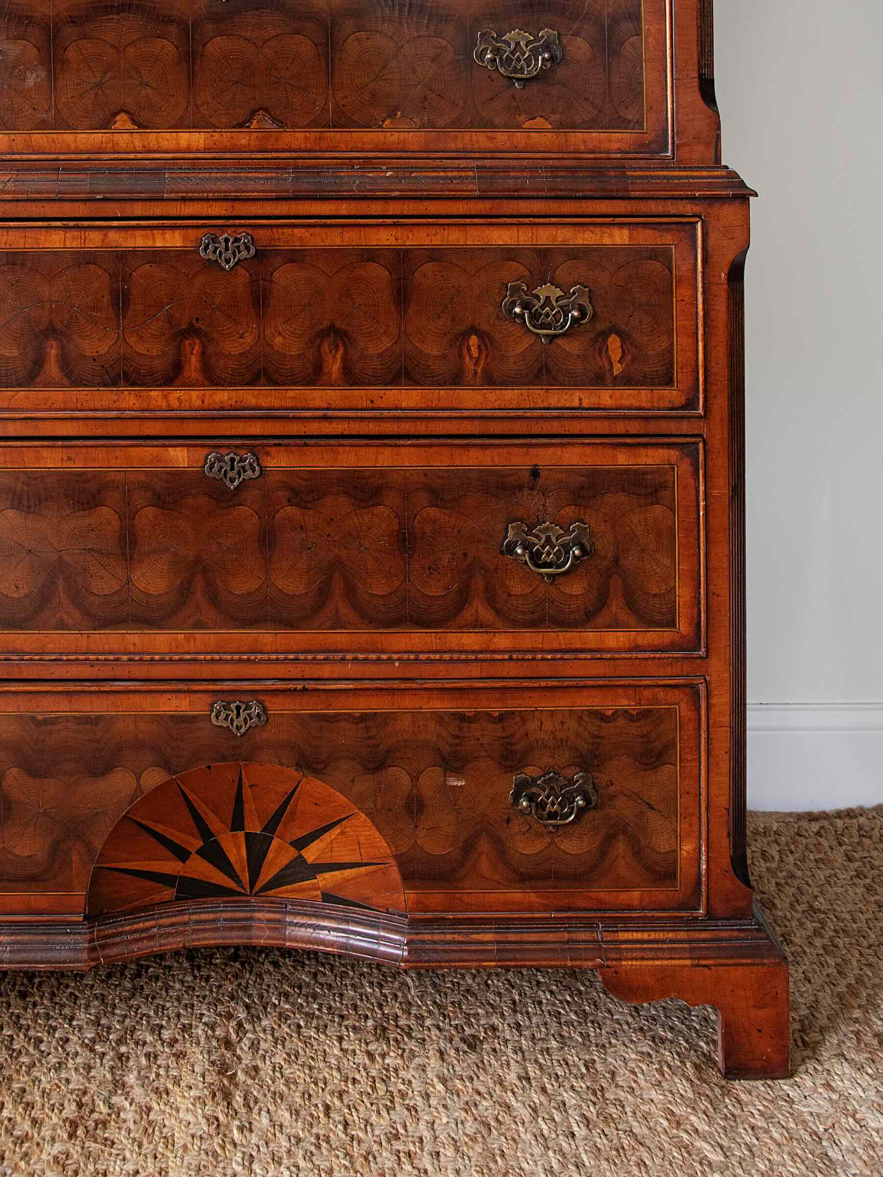 Close-up of an antique wooden chest of drawers with intricate marquetry patterns, brass drawer pulls, and a star inlay on the lower drawer, set on a woven rug against a white wall.