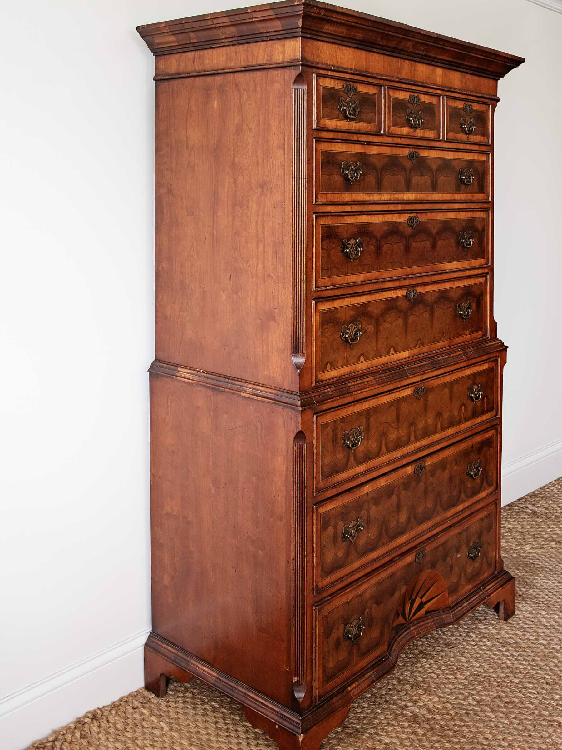 A tall, antique-style wooden chest of drawers with multiple drawers and ornate dark metal pulls, photographed in a room corner against a white wall and woven rug.