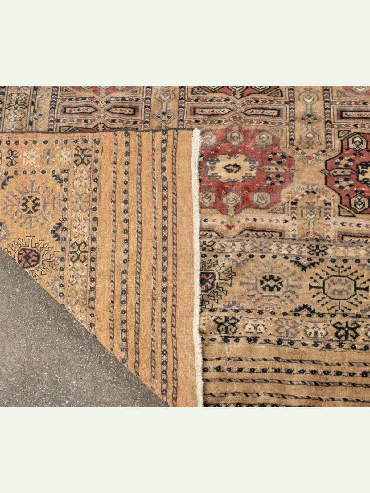 A close-up of a traditional patterned rug with a folded corner revealing the underside, showing beige, red, and black woven motifs on a light gray background.
