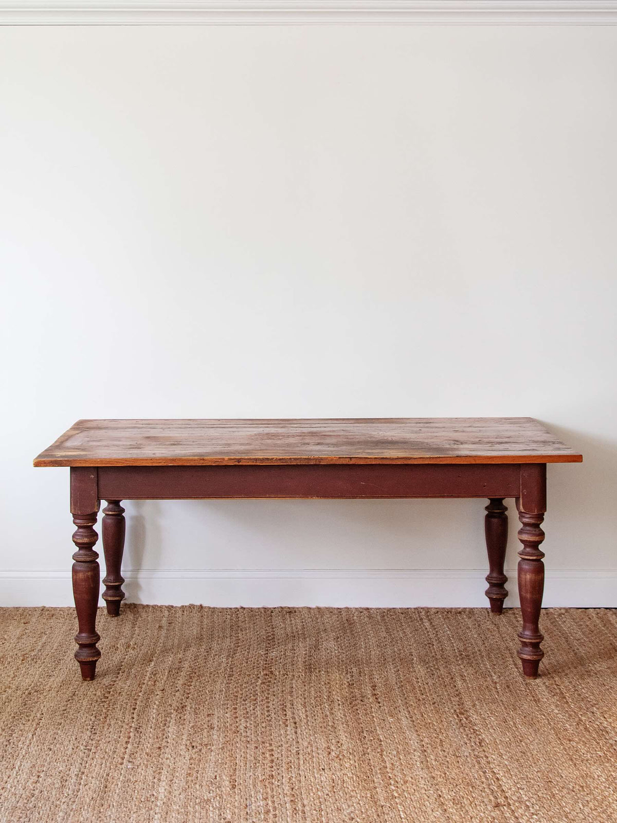 A rustic rectangular wooden table with turned legs, shown against a plain white wall on a woven jute rug.