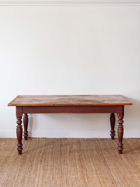 A rustic rectangular wooden table with turned legs, shown against a plain white wall on a woven jute rug.