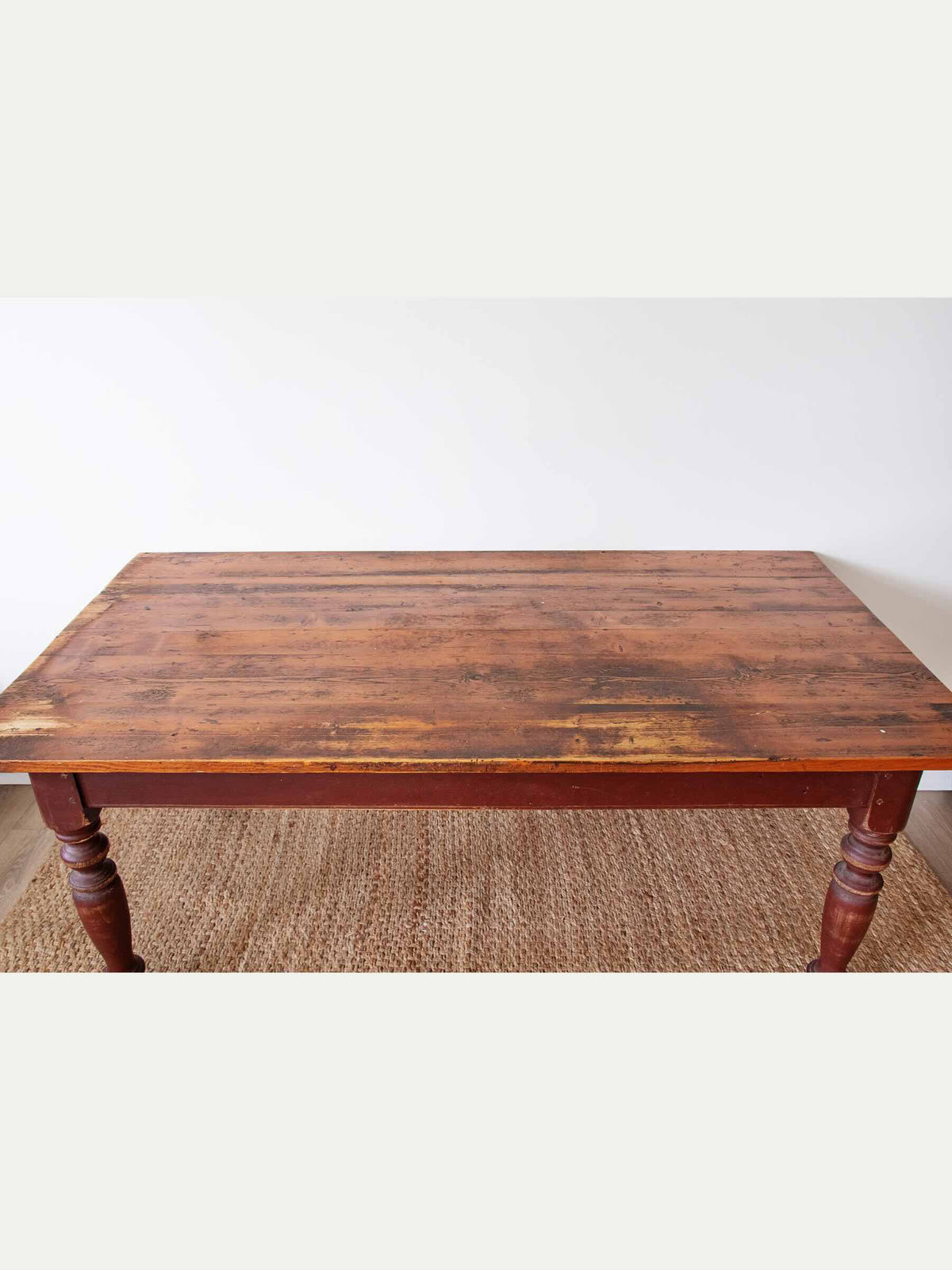 A rustic rectangular wooden table with a distressed brown finish and turned legs, shown on a woven jute rug against a plain white wall.