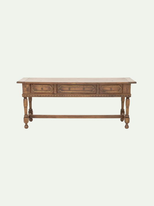 A rustic wooden console table with three drawers, turned legs, and a lower stretcher bar, shown against a solid off-white background.
