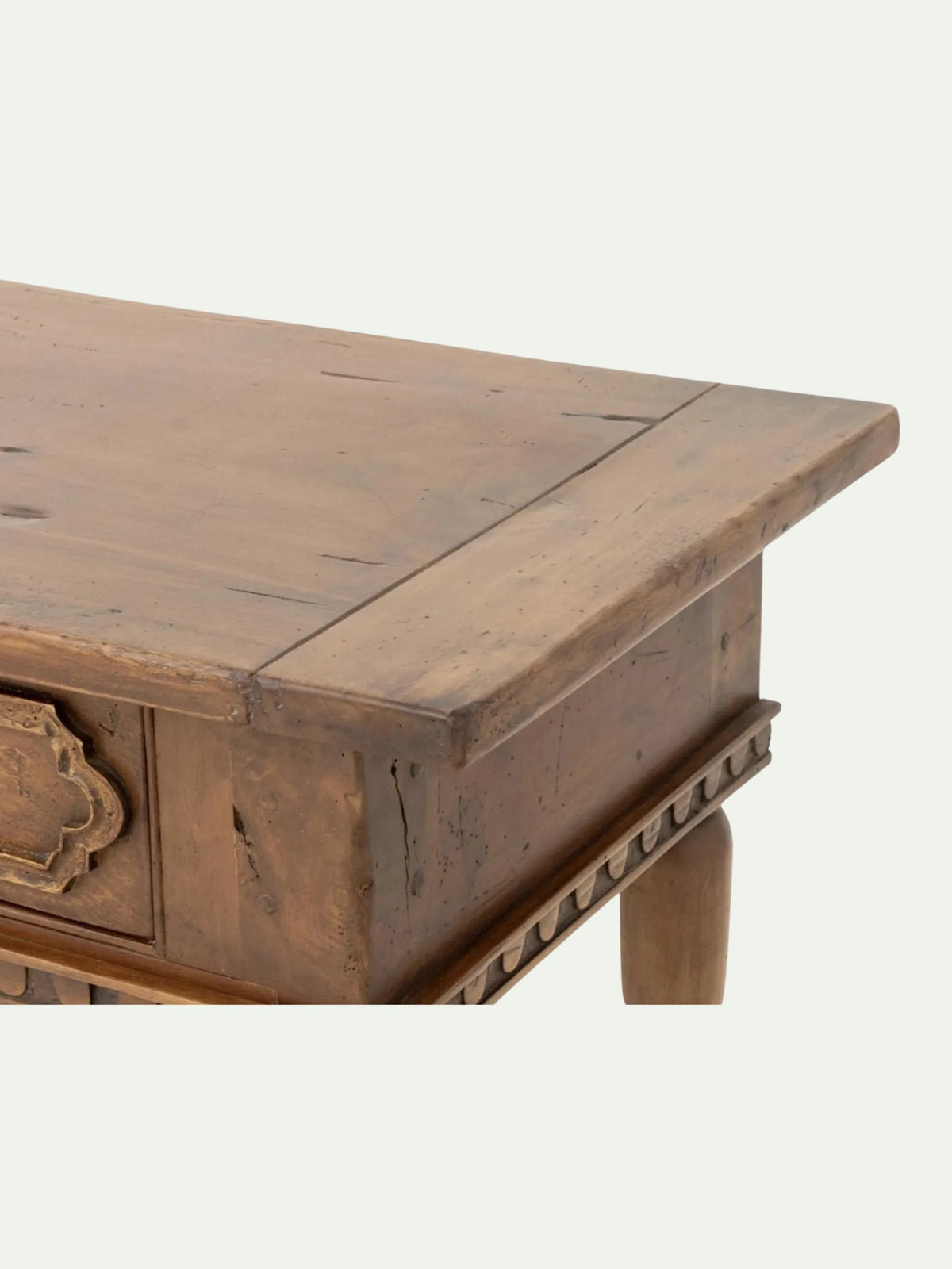 Close-up of a rustic wooden table corner showing the top surface, side panel, decorative drawer front, and leg against a plain light background.