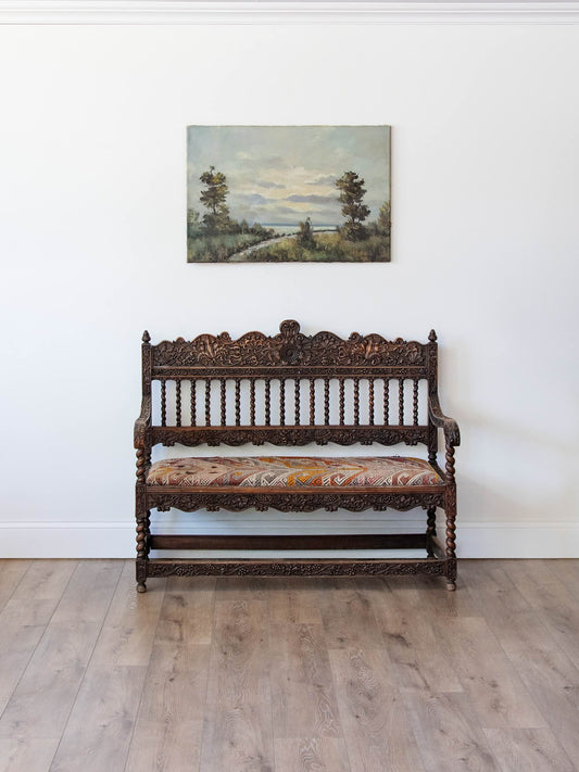 An ornate dark carved wooden bench with a patterned cushion sits against a white wall on a light wood floor, beneath a landscape painting.