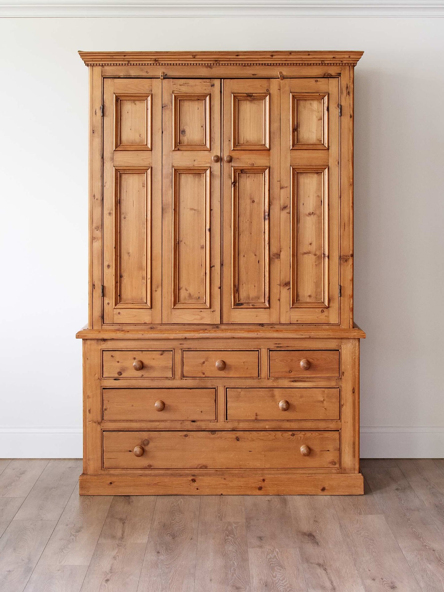 A large rustic pine wooden armoire cabinet with a six-panel upper door section and five drawers below, set against a plain white wall and light wood floor.