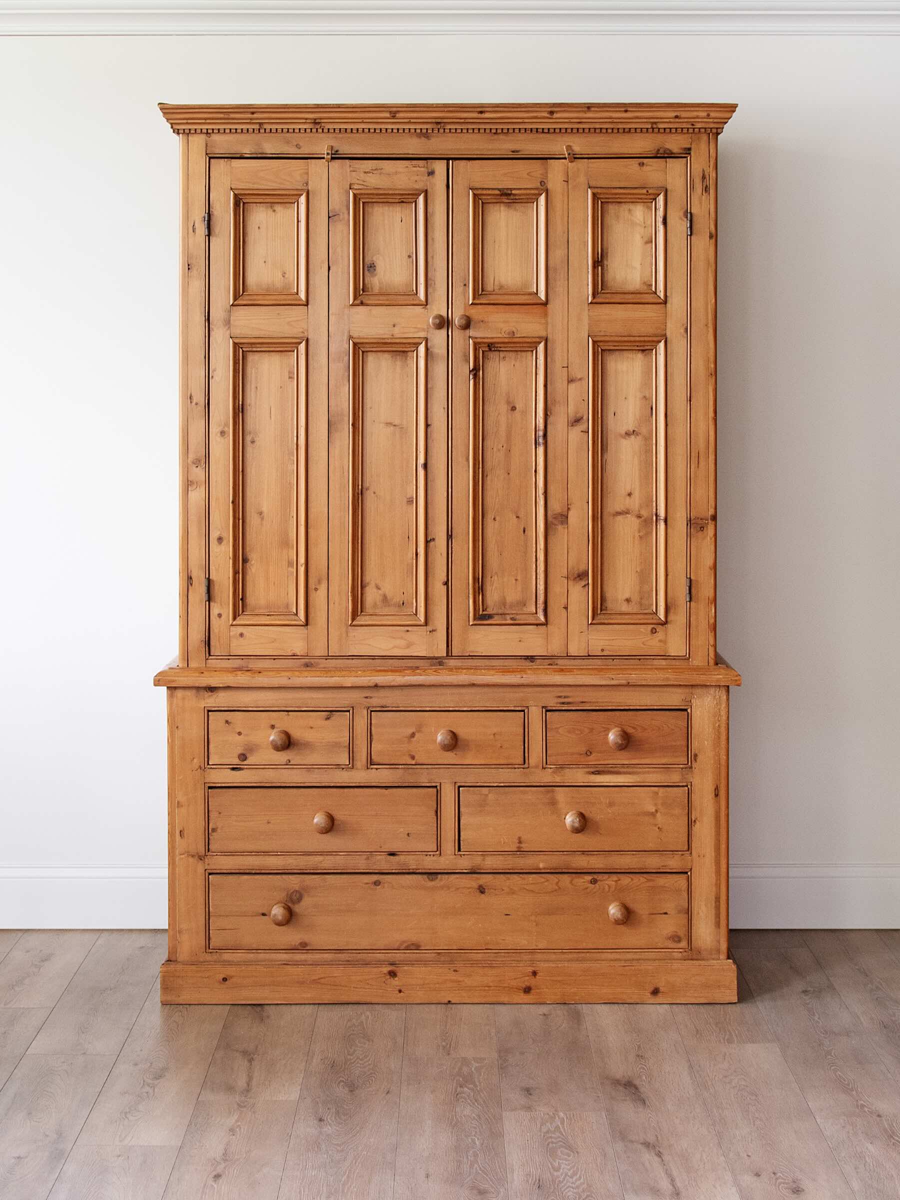 A large rustic pine wooden armoire cabinet with a six-panel upper door section and five drawers below, set against a plain white wall and light wood floor.