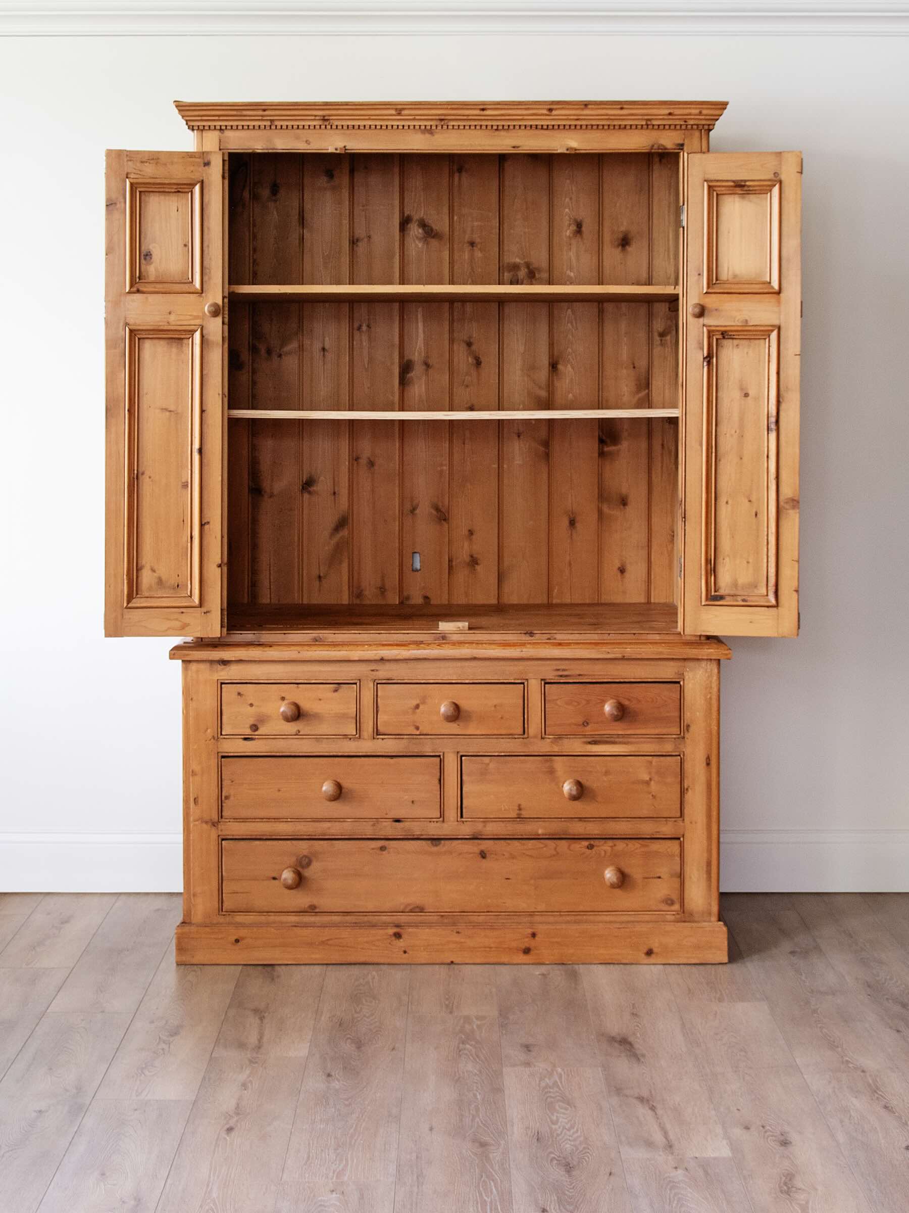 A rustic pine wooden hutch with an open door display, featuring a top shelf section with two doors and shelves, and a lower cabinet with five drawers, set against a white wall and light wood floor.