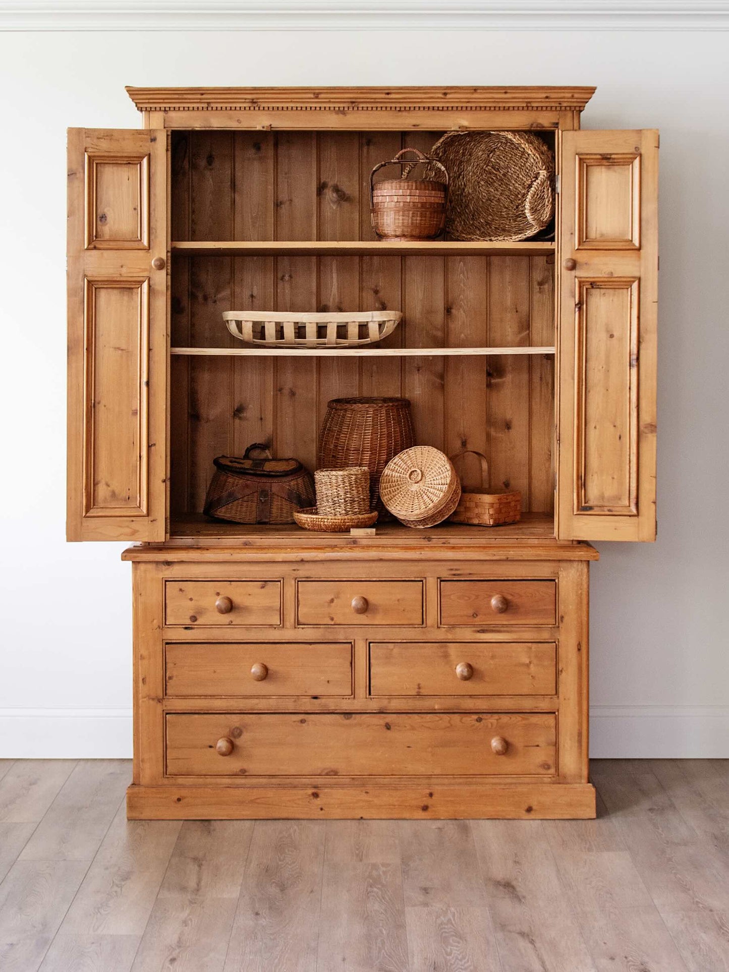 A rustic pine wooden hutch with open doors, featuring a lower drawer section and open shelves displaying woven baskets and decorative items, set against a white wall and light wood floor.