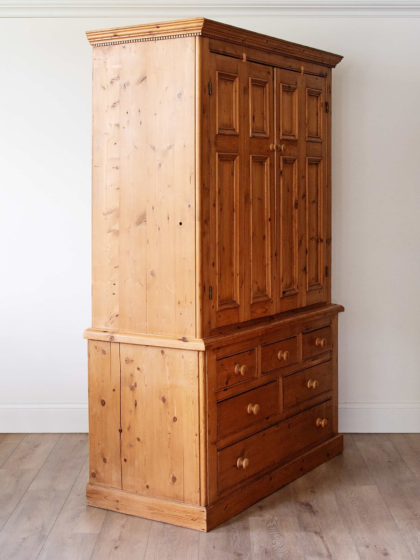 A tall wooden storage cabinet with a light pine lower section and a darker brown paneled upper section, shown in a room with white walls and light wood flooring.
