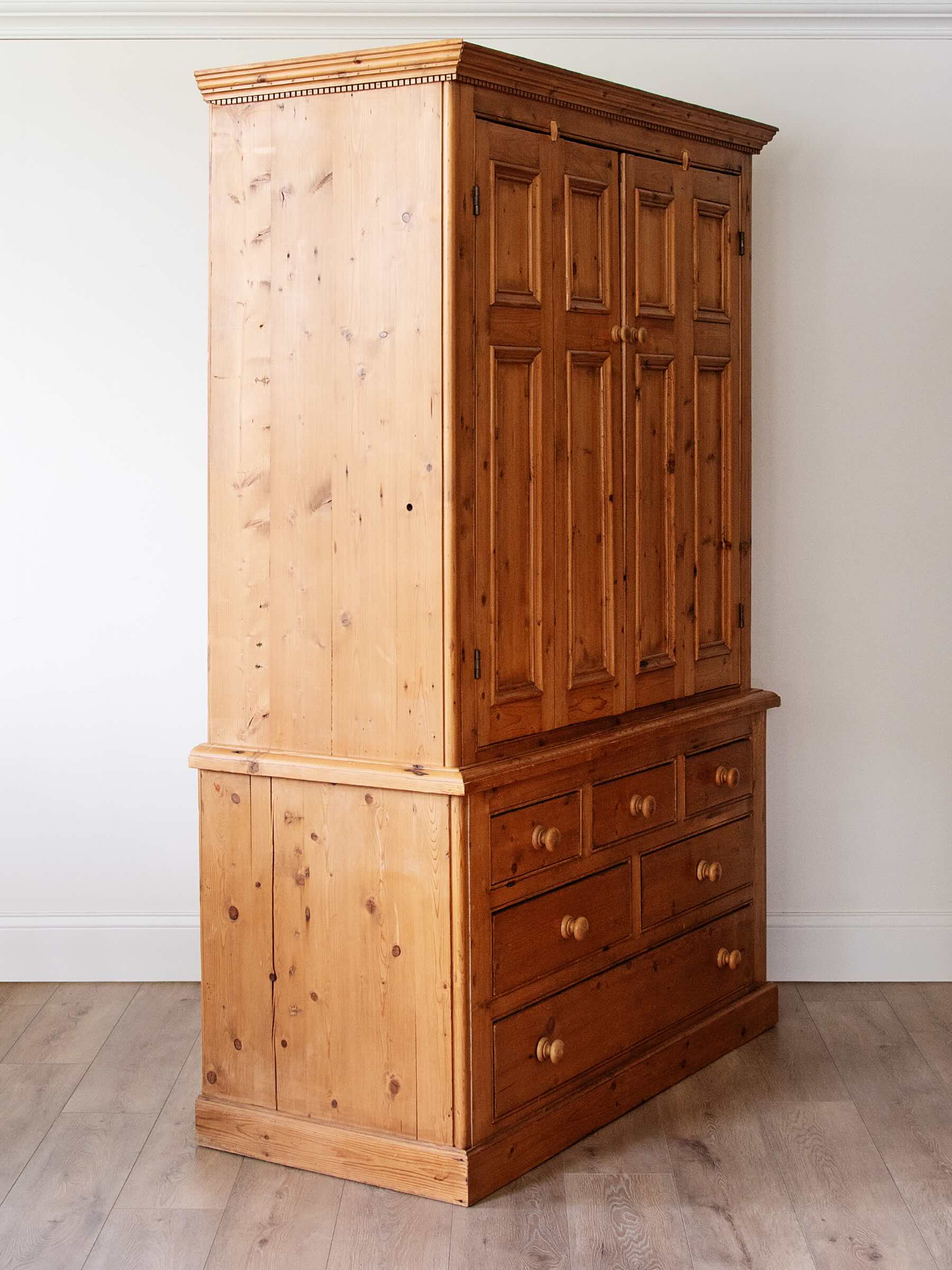 A tall wooden storage cabinet with a light pine lower section and a darker brown paneled upper section, shown in a room with white walls and light wood flooring.