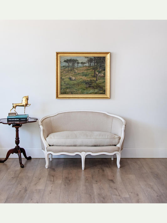 A classic French-style upholstered chaise lounge with a cream fabric and white carved frame, positioned in a room with light wood floors and a white wall. A gold-framed landscape painting is hanging on the wall above the lounge, and a small dark wood side table with decorative items is on the left.