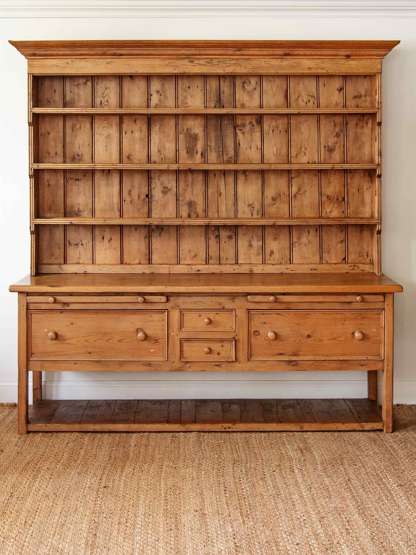 A rustic pine wooden hutch with a tall open shelving section on top, a work surface, three drawers, and an open lower shelf, shown against a white wall on a woven jute rug.
