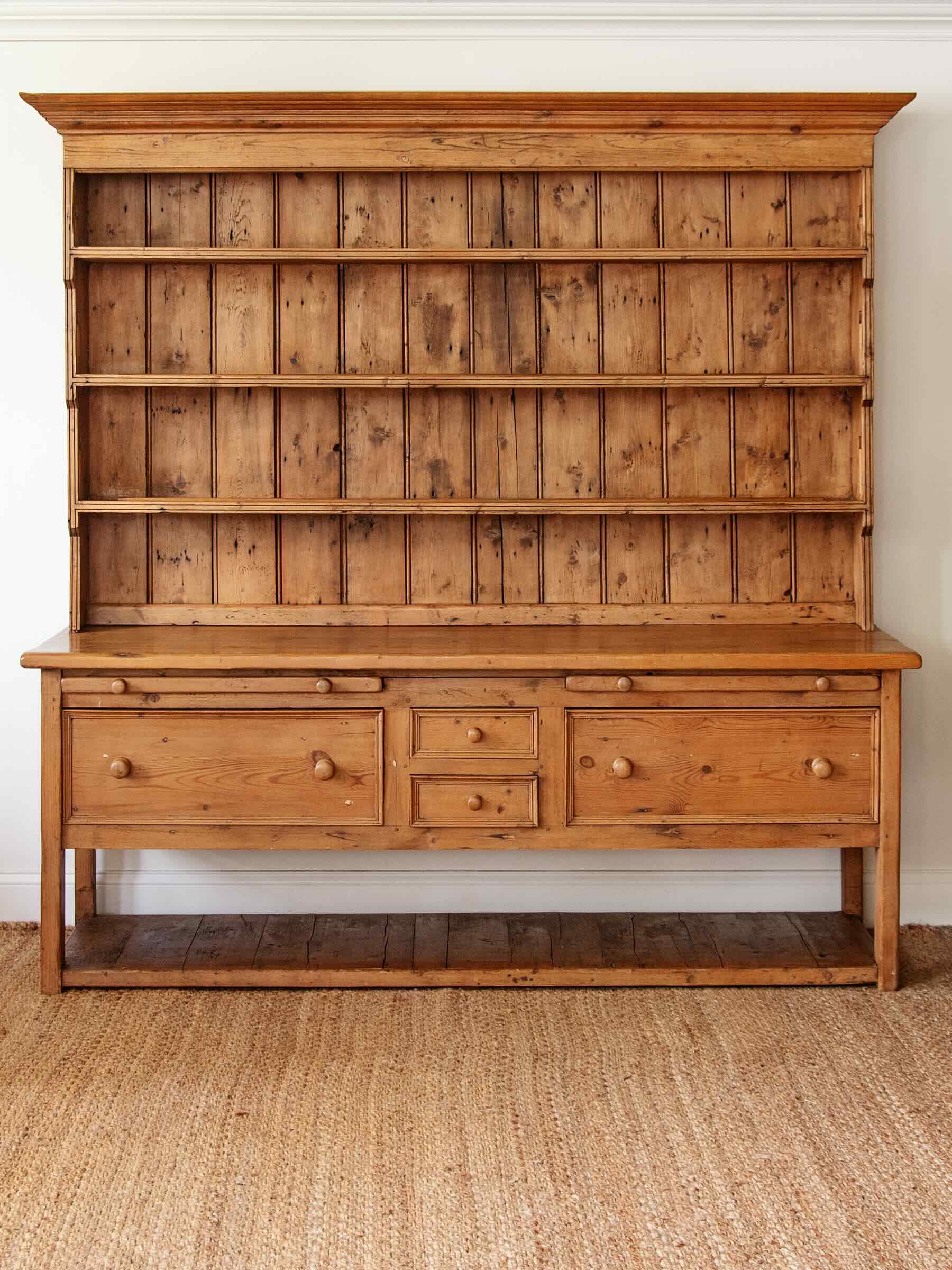 A rustic pine wooden hutch with a tall open shelving section on top, a work surface, three drawers, and an open lower shelf, shown against a white wall on a woven jute rug.
