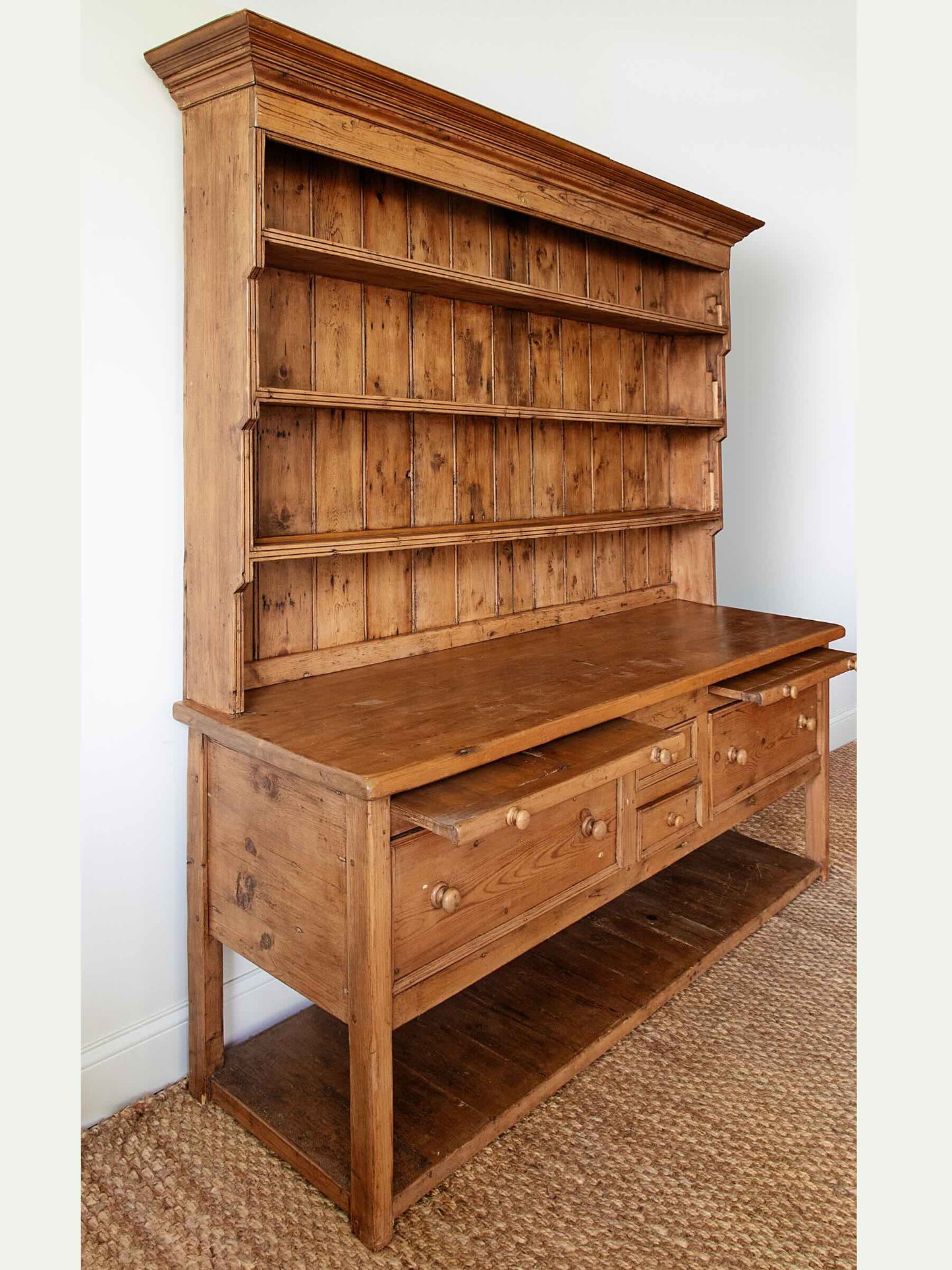 A rustic wooden hutch and bench combination piece with a tall open shelving back, a writing surface, three drawers, and an open lower shelf, shown in a room setting on a woven rug.