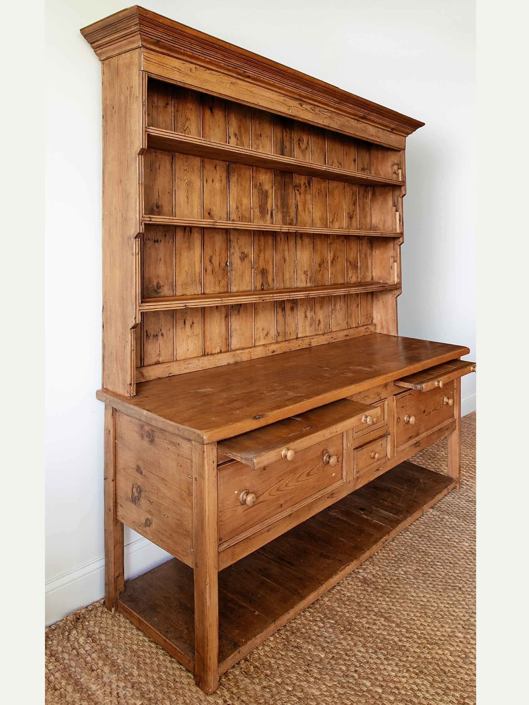 A rustic wooden hutch and bench combination piece with a tall open shelving back, a writing surface, three drawers, and an open lower shelf, shown in a room setting on a woven rug.