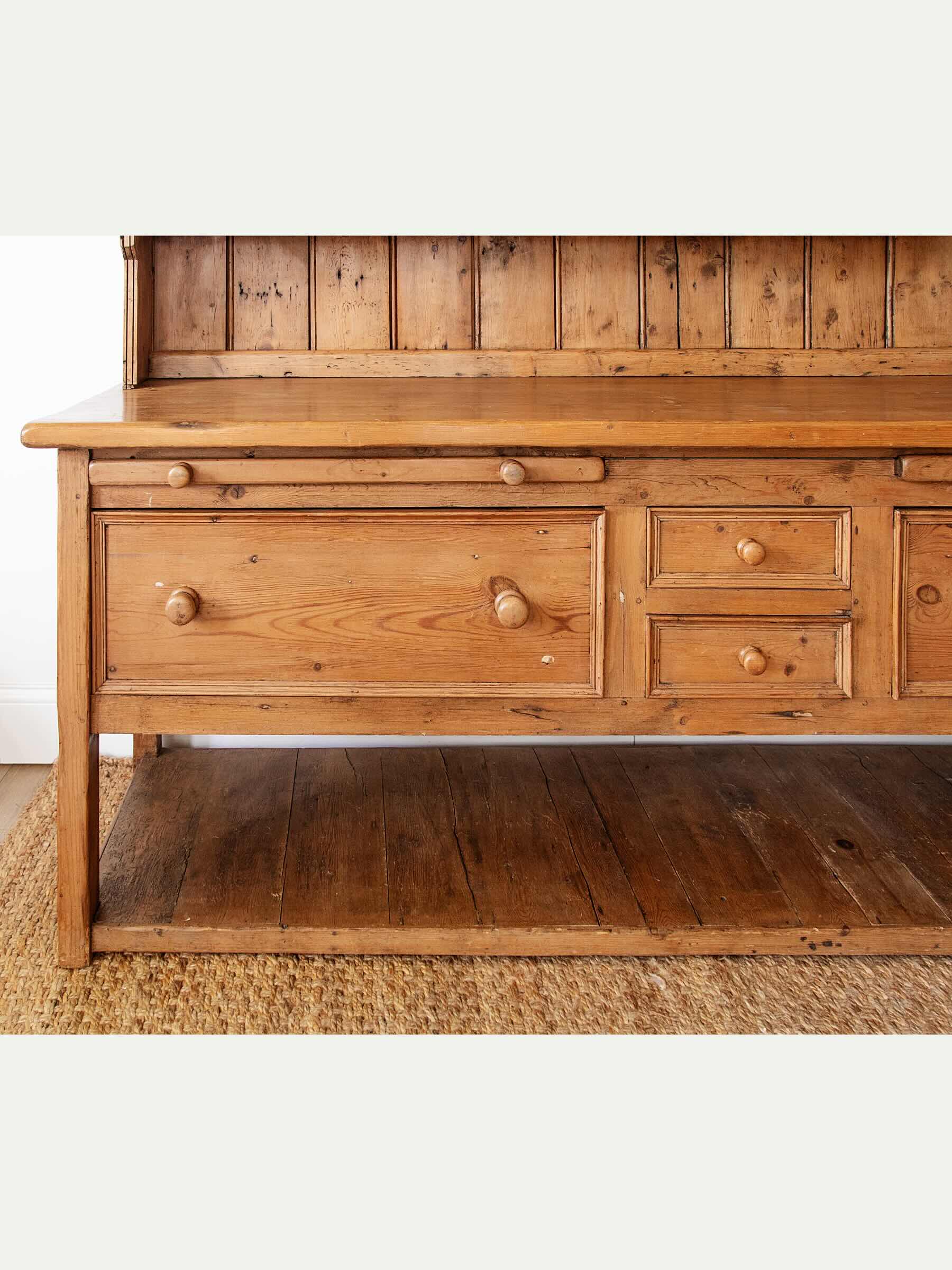 A rustic wooden hutch or chest of drawers with multiple drawers, round wooden knobs, a slatted back panel, and an open lower shelf, shown against a light neutral background.