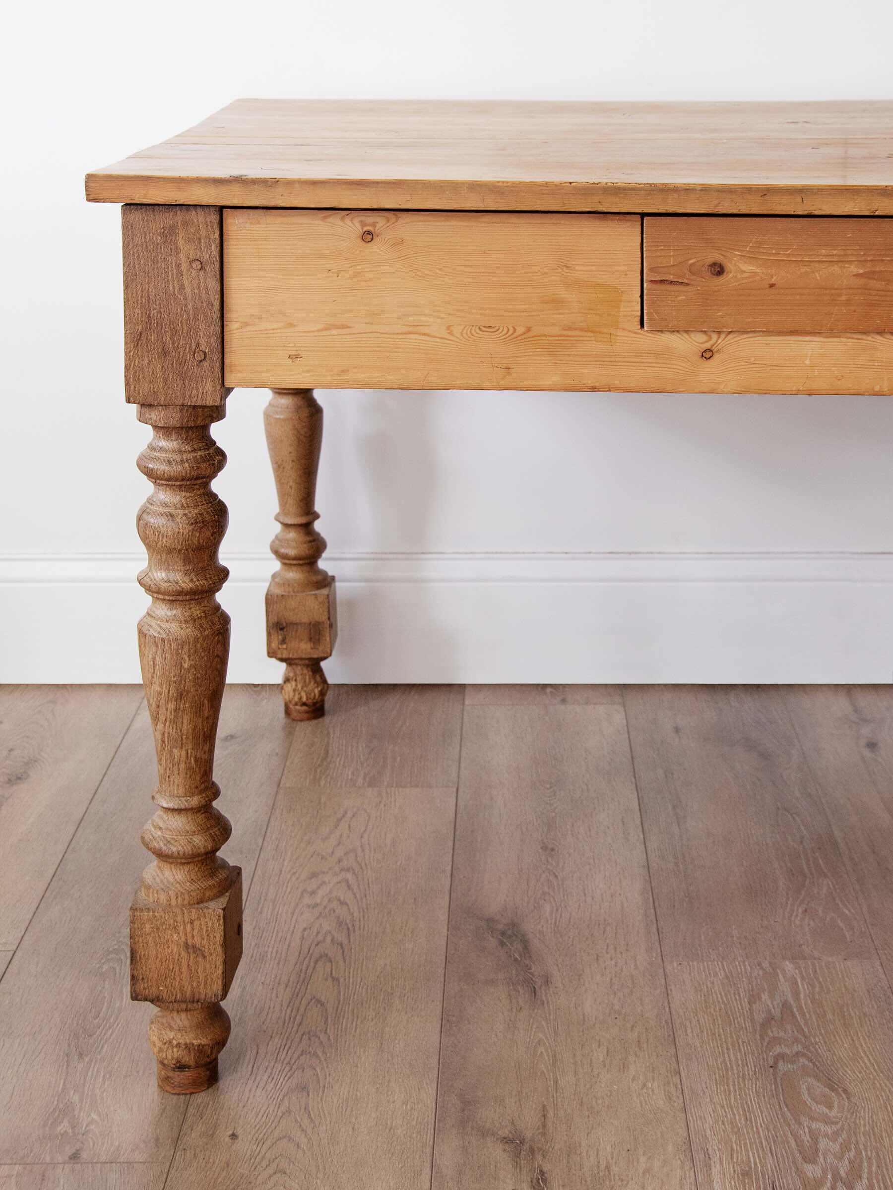 A rustic wooden writing desk with turned spindle legs and a single drawer, shown on a light wood floor against a white wall.