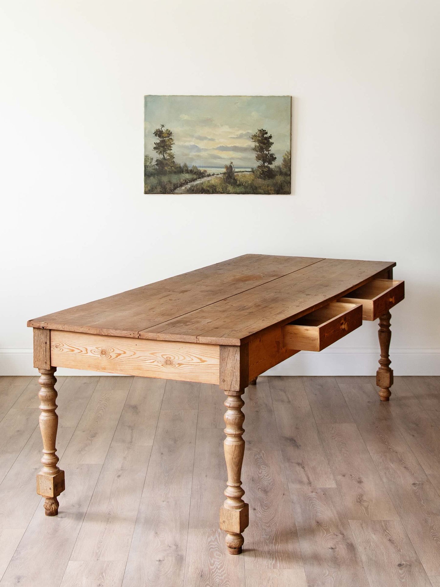 A rustic wooden writing desk with turned legs and two open drawers, placed on a light wood floor against a white wall with a landscape painting above.