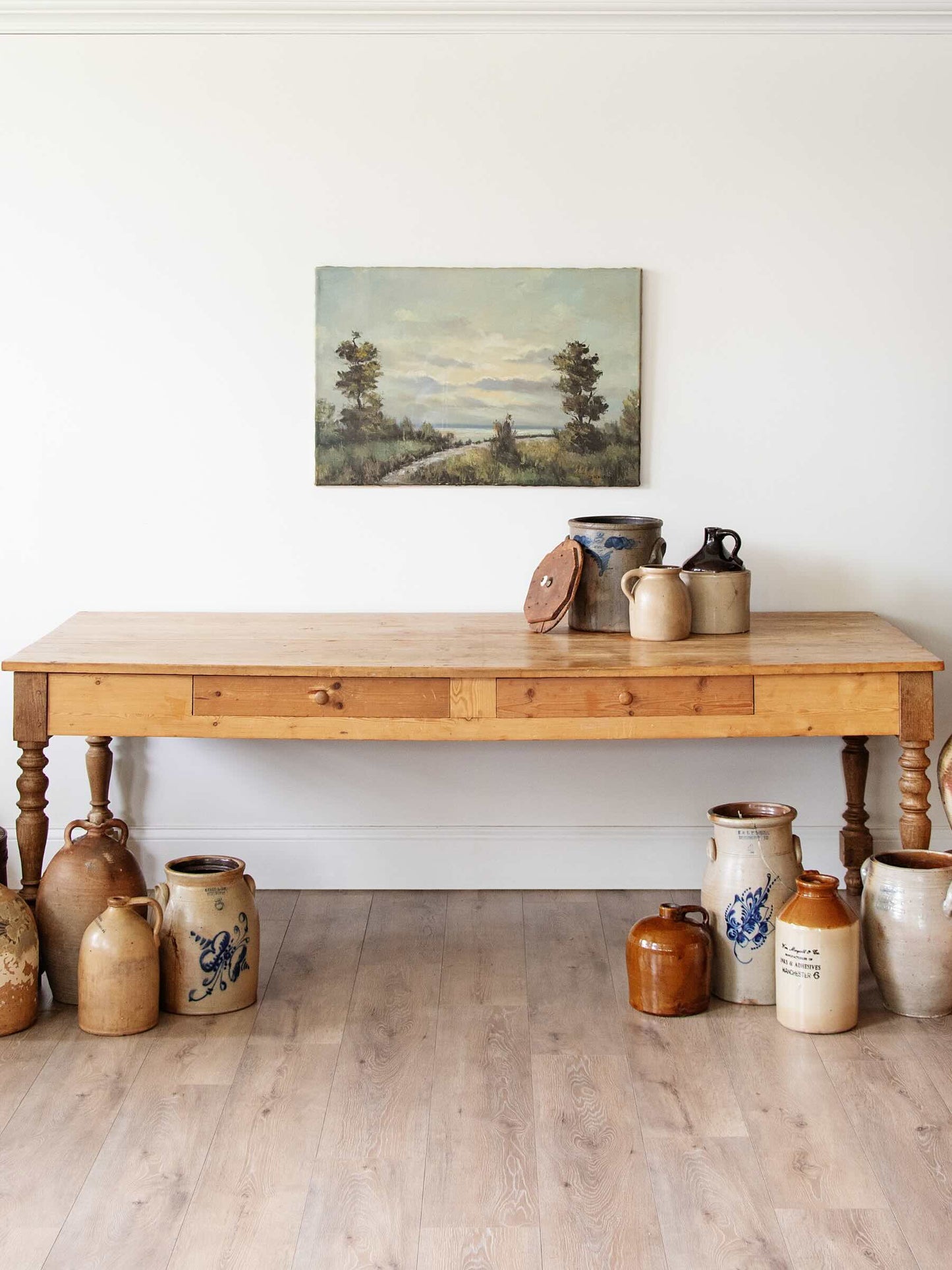 A rustic wooden console table with turned legs and drawer fronts, styled in a room with vintage stoneware jugs and a landscape painting on the wall above.
