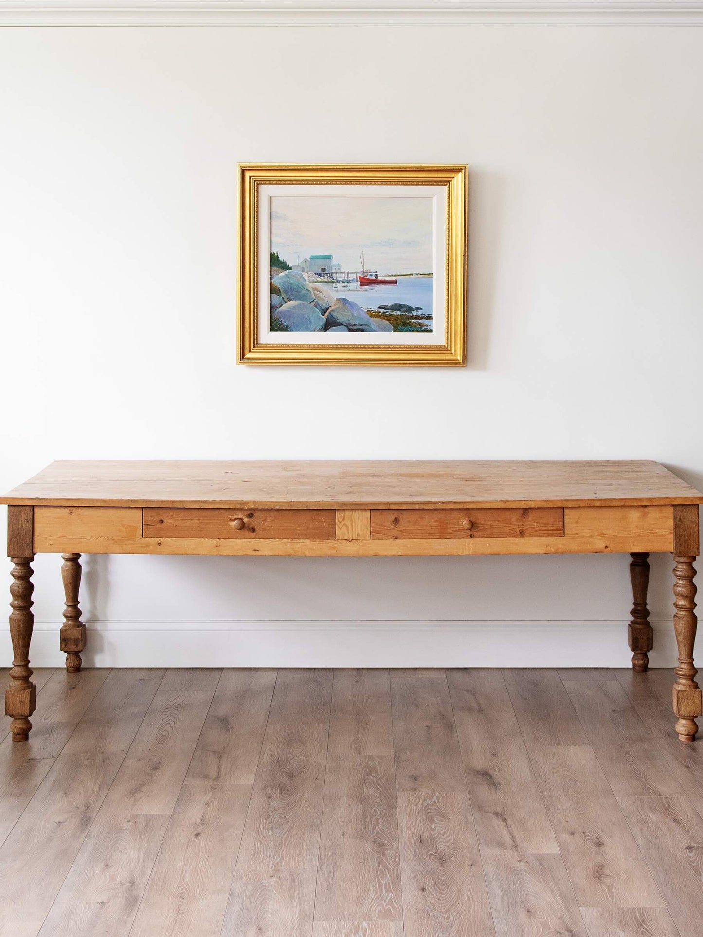 A rustic wooden console table with turned legs and a gold-framed coastal landscape painting hanging on a white wall above it, set against a light wood floor.