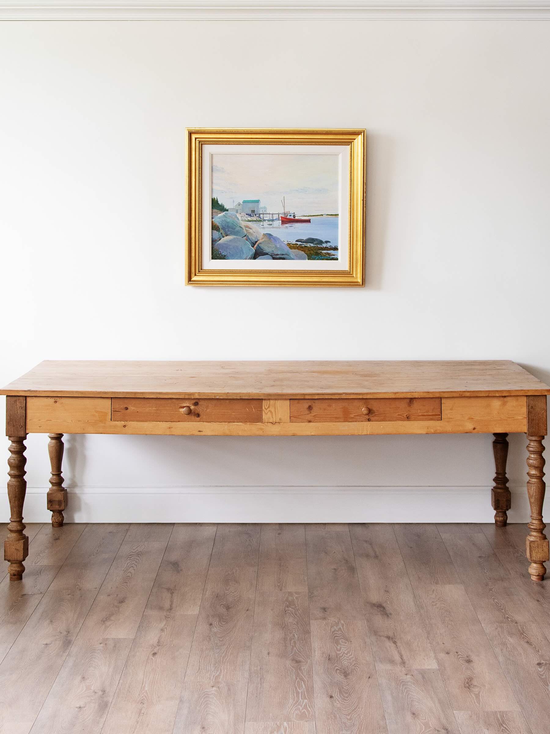 A rustic wooden console table with turned legs and a gold-framed coastal landscape painting hanging on a white wall above it, set against a light wood floor.