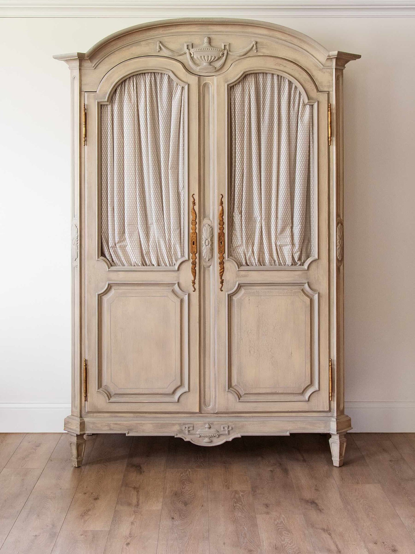 A large vintage-style painted wooden armoire cabinet with two arched door panels, draped cream curtains, ornate gold handles, and a distressed off-white finish, set against a plain wall and light wood floor.